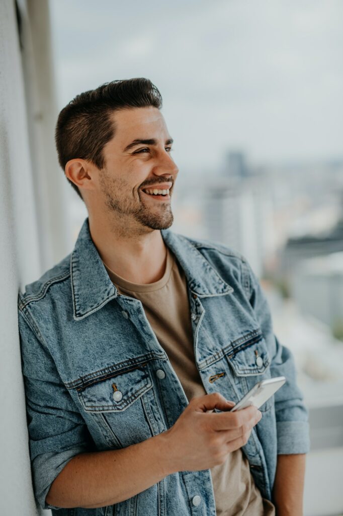 Portrait of happy young caucasian man with smartphone standing on balcony